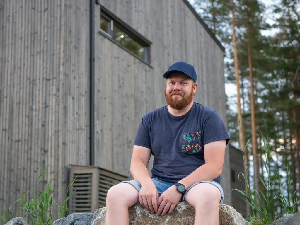 Jan Inge sittende på en stein utenfor huset sitt. Varmepumpe luft-til-luft i bakgrunnen.